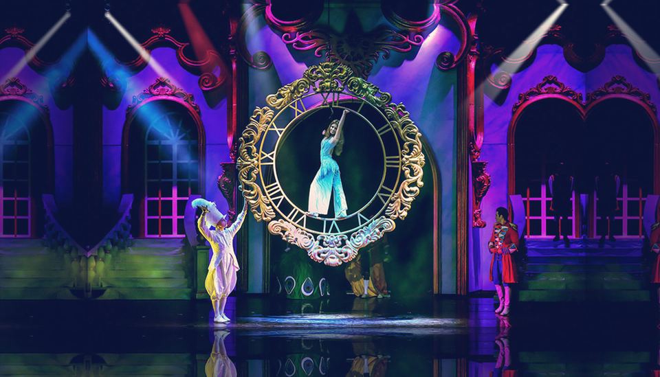 Escena de Cenerentola: una acróbata dentro de un gran reloj dorado, en un palacio iluminado en violeta.